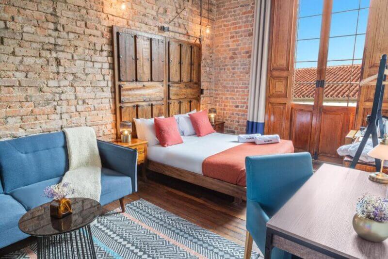 A cozy bedroom at Selina La Candelaria features exposed brick walls, a double bed with orange accents, a blue sofa, a desk, and large wooden doors that open to stunning views of rooftops and blue sky.