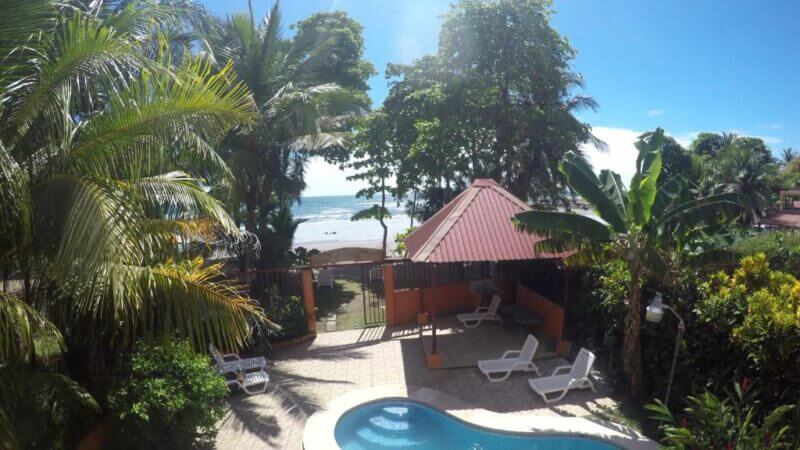 Experience La Dolce Vita with a view of a swimming pool, lounge chairs, and a gazebo nestled among tropical plants, with a gate opening to a sandy beach and the sparkling ocean beneath a clear blue sky.