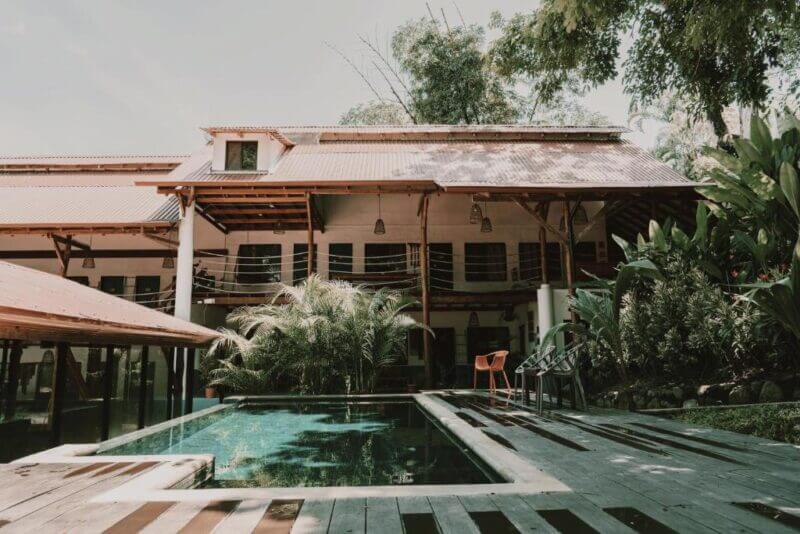 Two-story house with a red roof and wooden accents, surrounded by greenery, featuring a swimming pool and several chairs on a wooden deck at Selina Santa Teresa.