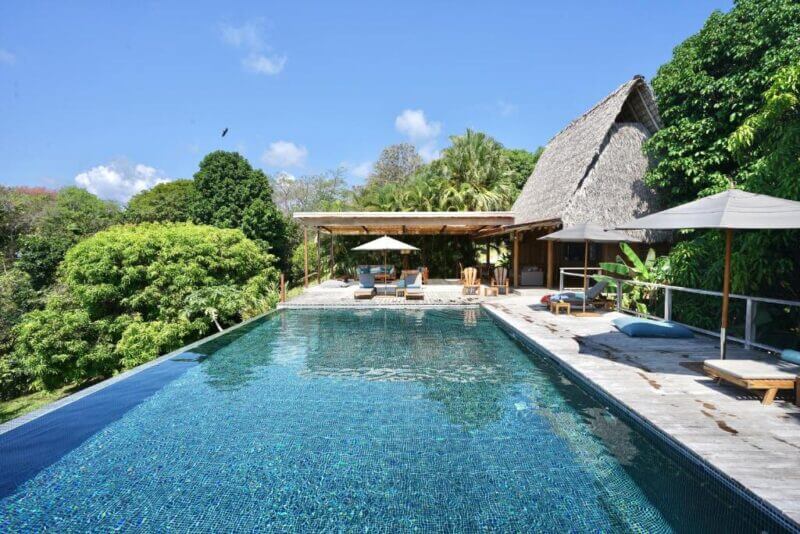 An infinity pool with clear blue water is surrounded by wooden decking, lounge chairs, and umbrellas at Calamocha Lodge, next to a thatched-roof building and lush tropical trees under a clear sky.