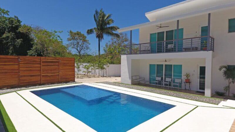 Modern two-story white house with balcony and covered patio, featuring a rectangular outdoor swimming pool and a wooden privacy fence, surrounded by trees and clear blue sky—perfect for your Nicoya Villas getaway.