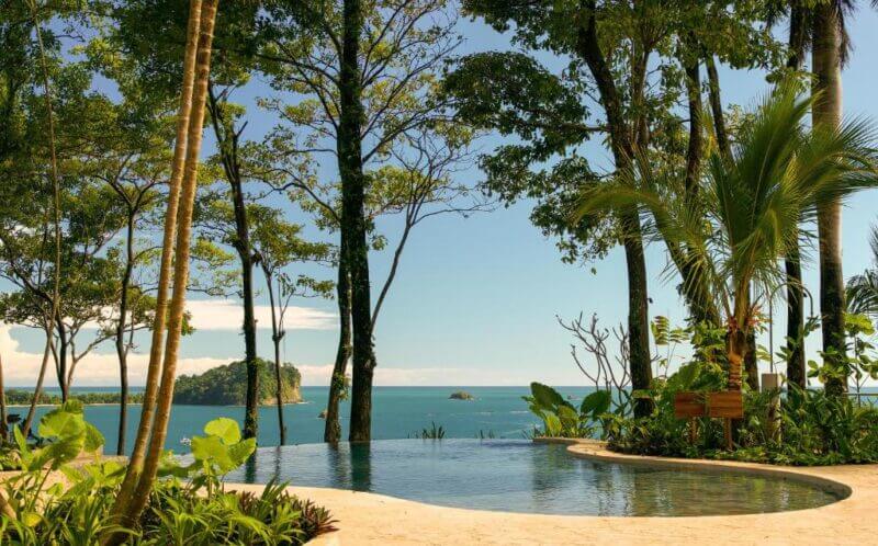 Infinity pool at Arenas Del Mar Resort, surrounded by tropical plants and trees, overlooking the ocean with two small islands visible in the distance under a clear sky.