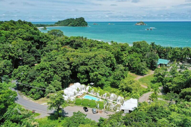 Aerial view of Igloo Beach Lodge, a coastal resort featuring white dome structures, a swimming pool, dense green trees, and the ocean in the background.