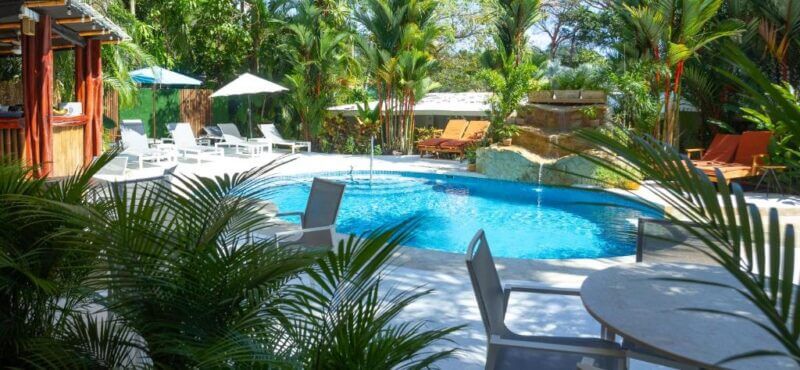 Outdoor pool area at La Vela Boutique Hotel with lounge chairs, tables, a small waterfall feature, palm trees, and a wooden bar surrounded by lush greenery.