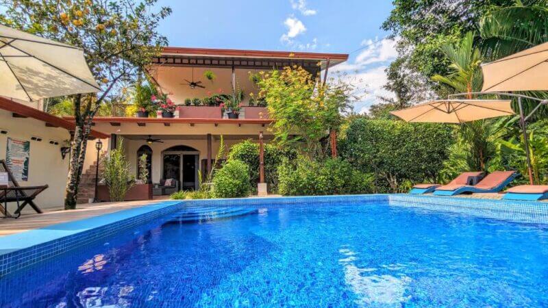 Two-story house with a balcony, nestled in the Jardín de los Monos, surrounded by lush greenery and viewed from a clear blue swimming pool with lounge chairs and umbrellas nearby.