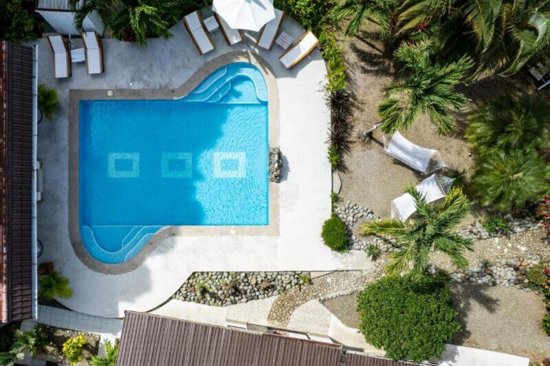 Aerial view of a rectangular swimming pool at Tribe Boutique Hotel, with lounge chairs, umbrellas, and surrounding greenery, next to a stone pathway and hammock area.