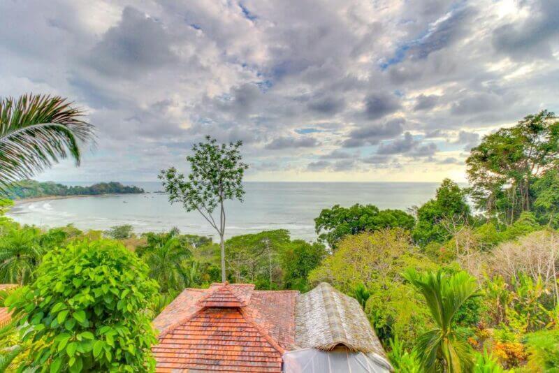 Ocean view with lush green trees and rooftops in the foreground under a cloudy sky. The shoreline curves gently along the bay, showcasing the scenic beauty of Canto del Mar in the background.