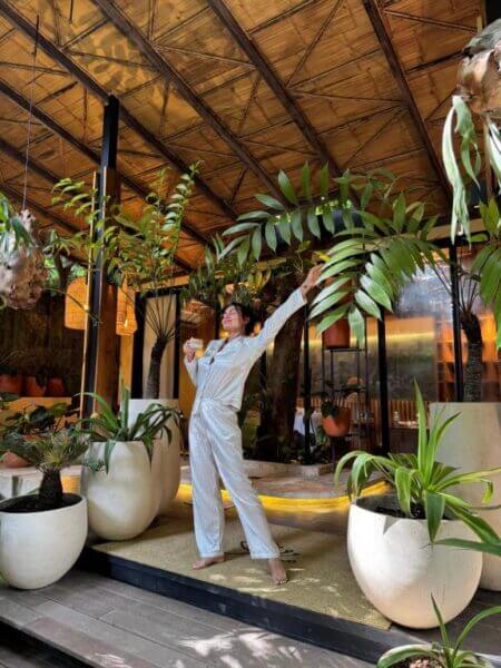 A person in striped pajamas stands on a patio at a boutique hotel, surrounded by large potted plants, holding a cup and stretching with one arm raised.