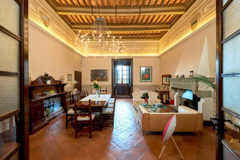 Spacious living and dining room at Il Tosco Montepulciano, featuring an ornate wooden ceiling, chandelier, artworks, a long dining table, fireplace, and a cozy seating area with armchairs and sofa.