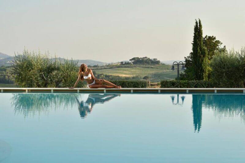 A woman in a white swimsuit sits at the edge of an outdoor pool at Borgobrufa Spa Resort, with hills and trees in the background.