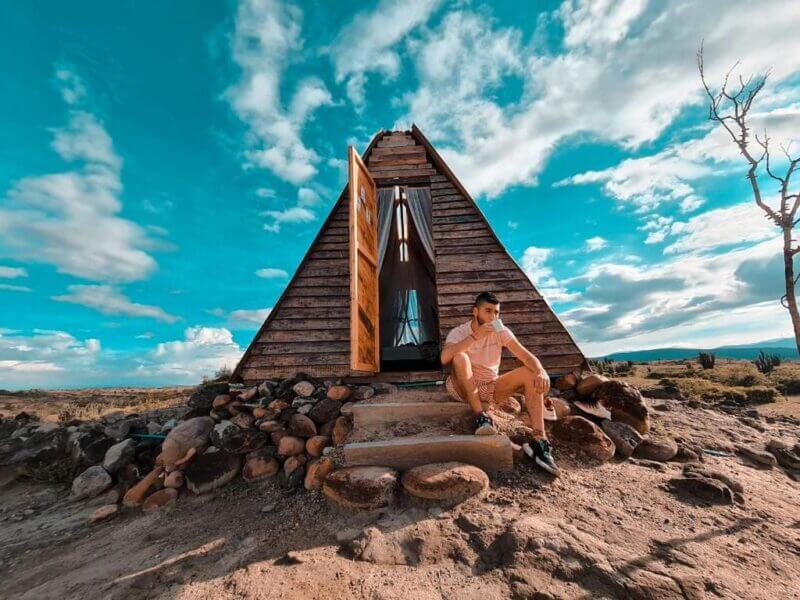 A person sits on the stone steps of a small A-frame wooden cabin at Mana Star Hotel, its door open to rocks and a natural landscape under a partly cloudy sky.