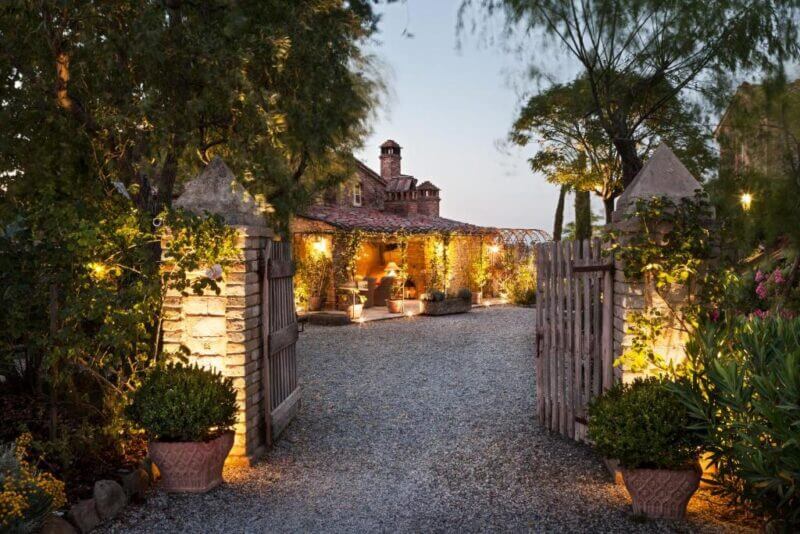 Stone gate opens to a gravel driveway leading to a rustic, warmly-lit house at Lupaia near Montepulciano, surrounded by trees and potted plants at dusk.