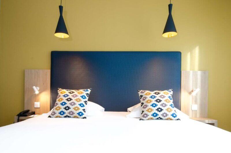 A neatly made bed with a blue headboard, two patterned pillows, and two black pendant lights against a yellow wall creates a cozy atmosphere at Best Western Villefrance.