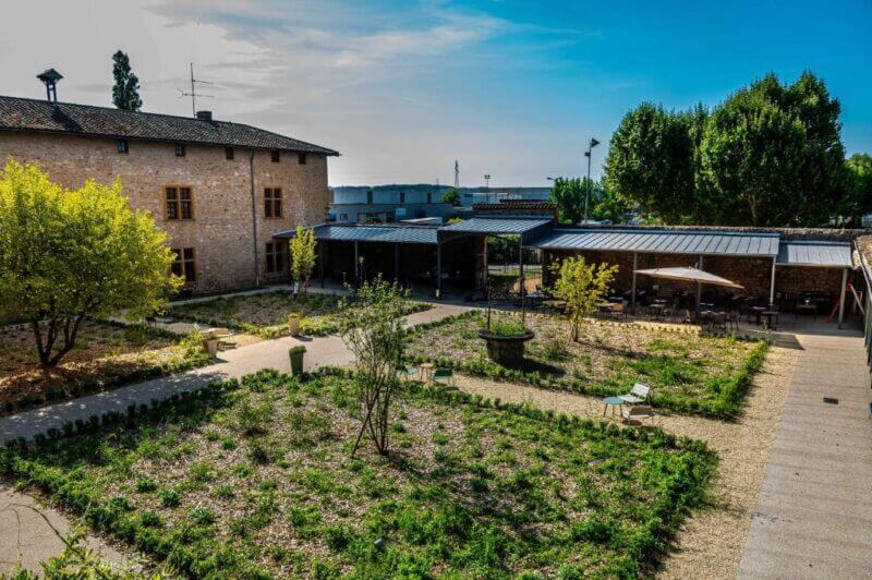 Courtyard garden at Hostellerie la Ferme du Poulet in Villefranche, featuring gravel paths, scattered plants, outdoor seating under canopies, and a stone building with wooden windows in the background.