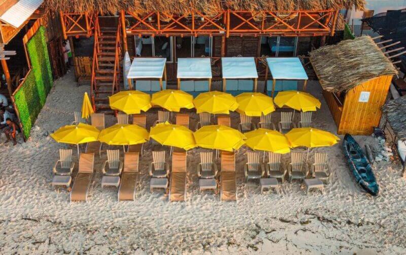 Aerial view of a beach setup with rows of lounge chairs under yellow umbrellas on sand, in front of a wooden beach hut and cabanas at Soy Local Barú. A blue kayak is placed nearby.