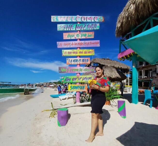 A woman stands smiling on a sandy beach near a colorful welcome sign and palm-thatched hut, with the vibrant BARU Bar in view, clear blue sky overhead, and people enjoying the lively background.