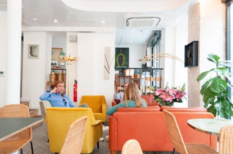 Two people sit on colorful armchairs in the bright, modern lounge at Hotel Lucien, surrounded by plants, flowers, and contemporary decor.