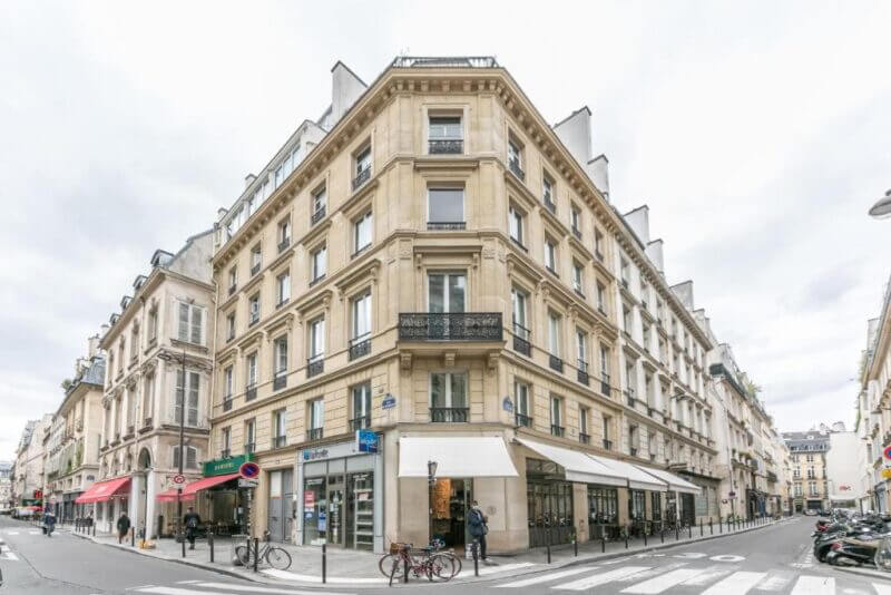 A beige, multi-story corner building—possibly Richelieu apartments—with shops at street level, balconies on upper floors, and bicycles parked along the sidewalk at a lively city intersection.