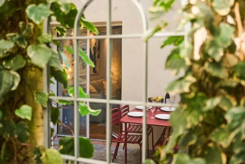 A red dining table with four chairs is set with plates and glasses in a cozy indoor space, viewed through a window framed by leafy plants—offering a glimpse of Boutik Boheme's inviting Le jardin ambiance.
