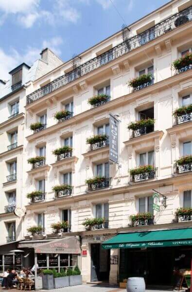 Four-story cream-colored boutique hotel with flower boxes on each window, a vertical Grand Hôtel Lévêque sign, and shops with green and brown awnings at street level on a sunny day.