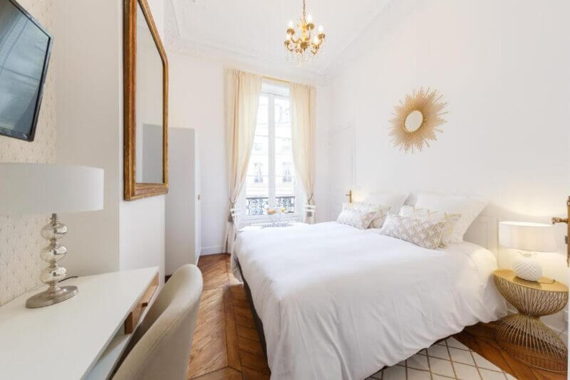 Bright bedroom with white walls, a large bed, decorative pillows, a chandelier, a wall mirror, and a window with sheer curtains allowing natural light—an inviting retreat inspired by Maison de Lignières elegance.