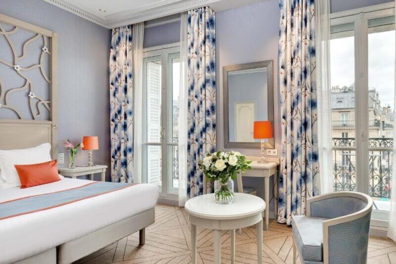Bright Splendid Etoile hotel room with a double bed, patterned curtains, two windows, a small table with flowers, a desk with a mirror, and a city view through the windows.