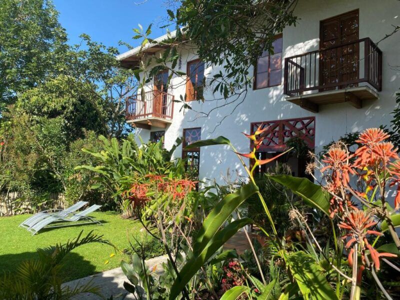 A two-story white Salento hotel with wooden balconies overlooks a garden filled with lush green plants and orange flowers under a clear blue sky.