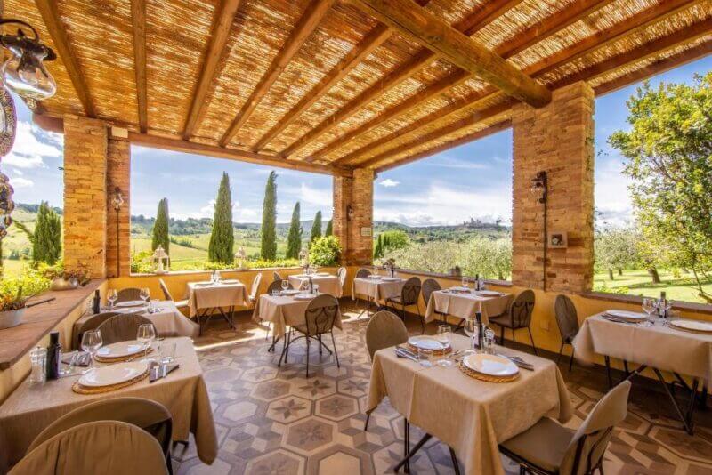 Outdoor restaurant terrace at Il Segreto di Pietrafitta Agriturismo, with tables set for dining and stunning views of the scenic countryside, lush trees, and greenery under a clear, sunny sky.