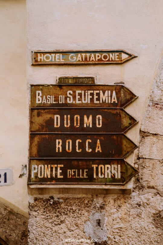 Weathered directional signs on a stone wall point toward Hotel Gattapone, Basil di S. Eufemia, Duomo, Rocca, and Ponte delle Torri—highlighting classic things to do in Umbria.