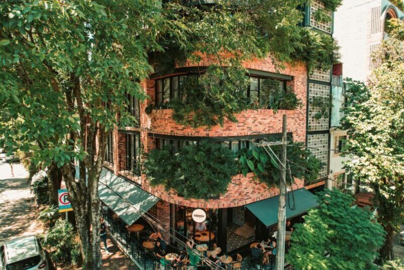 A red brick building with large windows and abundant greenery on its facade, the Nomada Hotel Medellin features an inviting outdoor seating area at street level under the shade of trees.