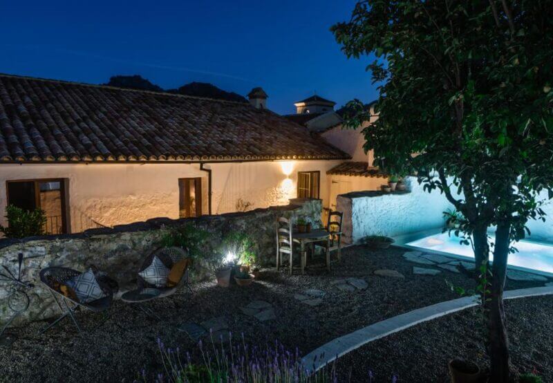 Courtyard at night with stone path, outdoor seating, potted plants, a small table with chairs, and a lit swimming pool next to Casa De Los Cuadros, a rustic house with a tiled roof.