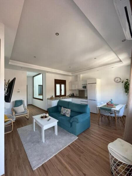 Modern open-plan living room and kitchen at Apartamentos Cantarería Jazmín, featuring wooden flooring, a teal sofa, white coffee table, dining area, and neutral decor.