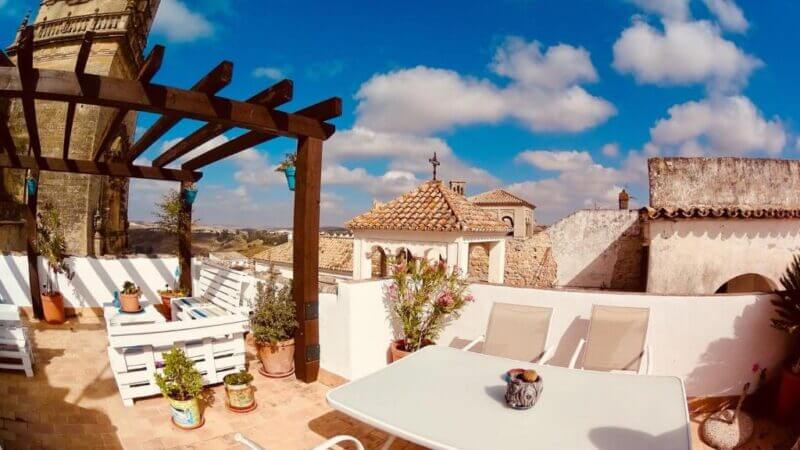 Sunny rooftop terrace at Casa Mirador with white outdoor furniture, potted plants, and a pergola, overlooking the old stone buildings of San Pedro under a blue sky with scattered clouds.
