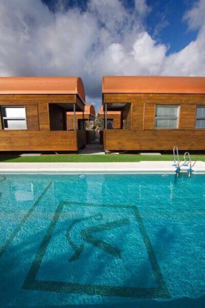 A clear swimming pool with a diving symbol at the bottom sits in front of two modern wooden buildings with orange roofs, nestled beside the Vía Verde and framed by the scenic Sierra under a partly cloudy sky.