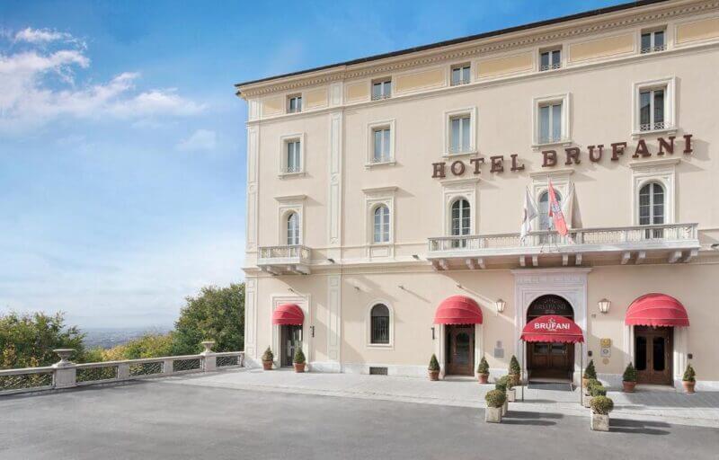 Exterior view of Sina Brufani, a luxury Perugia hotel housed in a historic cream-colored building with red awnings, several potted plants, and three flags at the entrance.