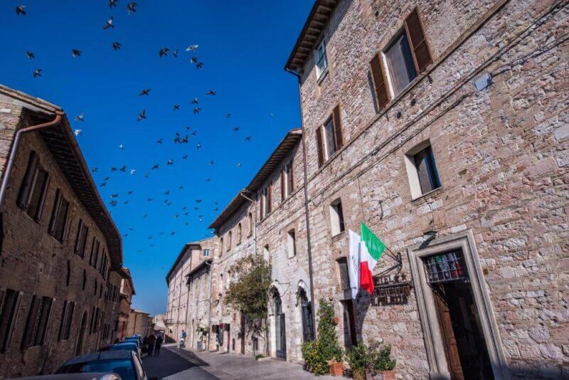 Stone buildings line a narrow street under a clear blue sky, with a flock of birds flying overhead and the Italian flag hanging from Hotel Il Palazzo Assisi.