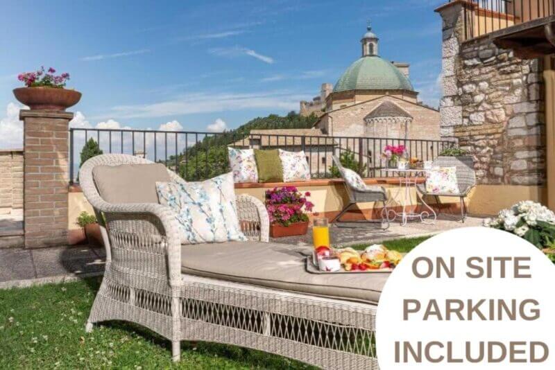 Outdoor terrace at Hotel Ideala Assisi with wicker lounge chairs, table set for breakfast, potted plants, and a historic dome in the background. Text reads: ON SITE PARKING INCLUDED.