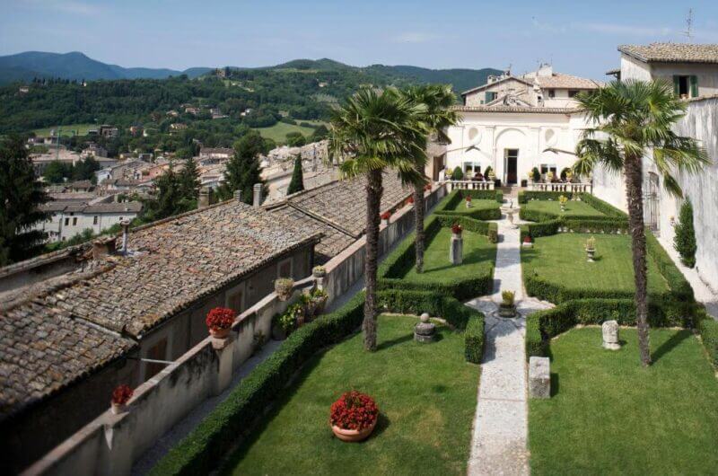 A formal garden with manicured hedges, stone path, potted plants, and statues at Palazzo Leti is bordered by historic buildings in Spoleto and overlooks a hilly landscape under a clear sky.