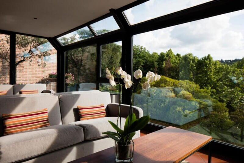 A modern living room with grey sofas, striped cushions, a Duchi wooden coffee table with a white orchid, and large glass windows overlooking green trees.