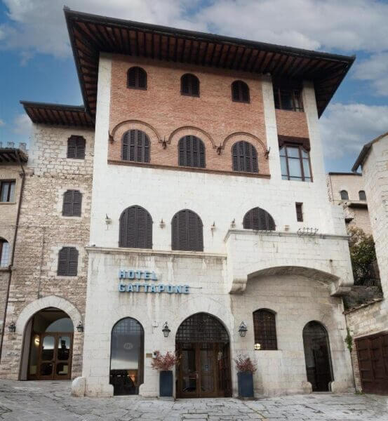 A stone and brick hotel building in Gubbio with arched doorways and shuttered windows, featuring a sign that reads Hotel Gattapone.