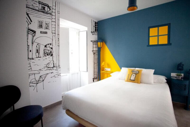 A modern bedroom in boutique hotel style features a white bed, cartoon-style wall art, a blue accent wall with a yellow window design, and a yellow pillow—perfect for unique Seville accommodation at Lukanda Seville.