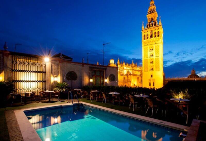 Rooftop pool and seating area at night at Doña Maria, with the illuminated Giralda tower and Seville Cathedral in the background.