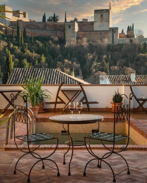 A small patio at Casa Bombo with two empty chairs and a table set with wine glasses, overlooking the historic fortress of Granada on the hill in the background.