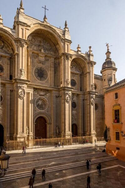 The image shows the ornate stone facade of Casa de la Catedral in Granada, featuring arched doorways, round windows, statues, and people walking on the steps below.