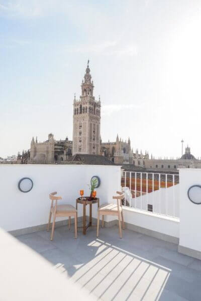 A rooftop terrace with two wooden chairs and a small table overlooks the Catedral and Giralda tower, offering stunning views of Sevilla under a clear sky.