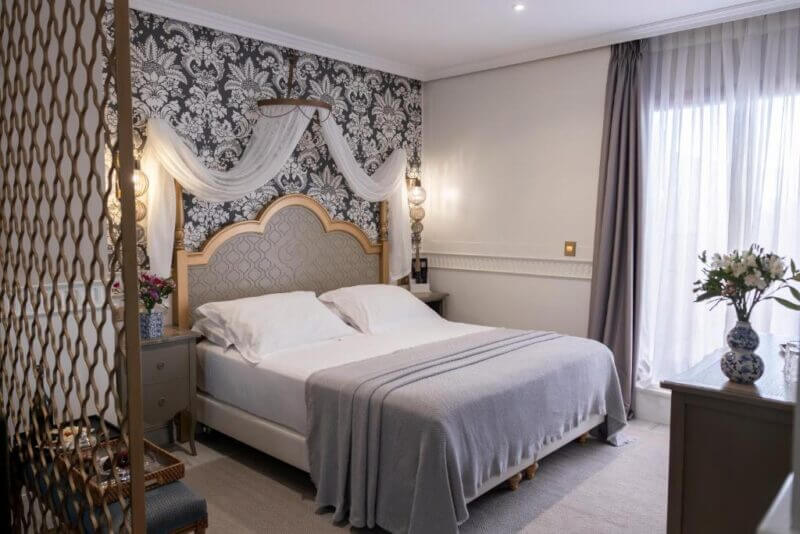 A neatly made double bed with gray bedding sits against a patterned accent wall, flanked by nightstands and lamps, in a softly lit, elegant bedroom with a window and sheer curtains at Hotel Gravina 51 in Seville.