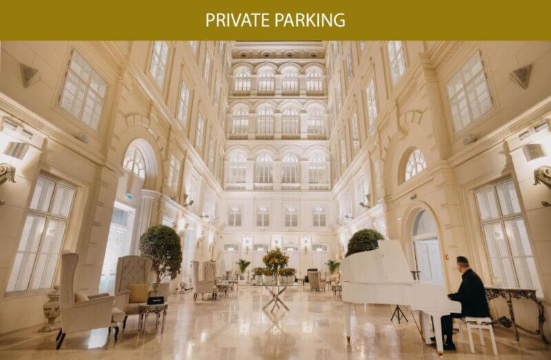 Elegant hotel atrium with high ceilings, ornate decor, large windows, and a person playing a white grand piano; Barcell Brno PRIVATE PARKING text appears at the top.