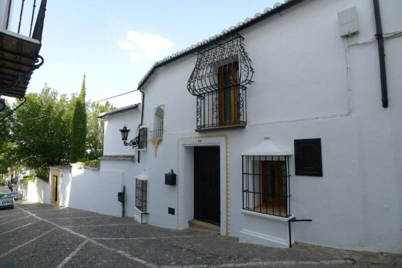 White, two-story Salvatierra Guest House with barred windows and a black door, set on a sloped cobblestone street and bordered by a white wall and trees in the background.