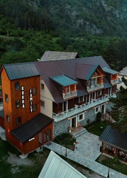 A multi-story wooden and stone Gurra Family Guesthouse with balconies, surrounded by greenery and mountains, featuring a welcoming sign reading Gurra Family.
