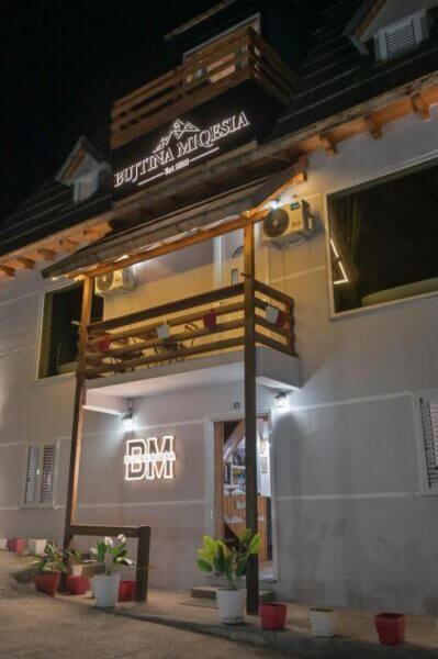 Night view of the two-story Bujtina Miquesia guesthouse with wooden balconies, potted plants at the entrance, and illuminated signs reading Miquesia Hotel and BM on the facade.